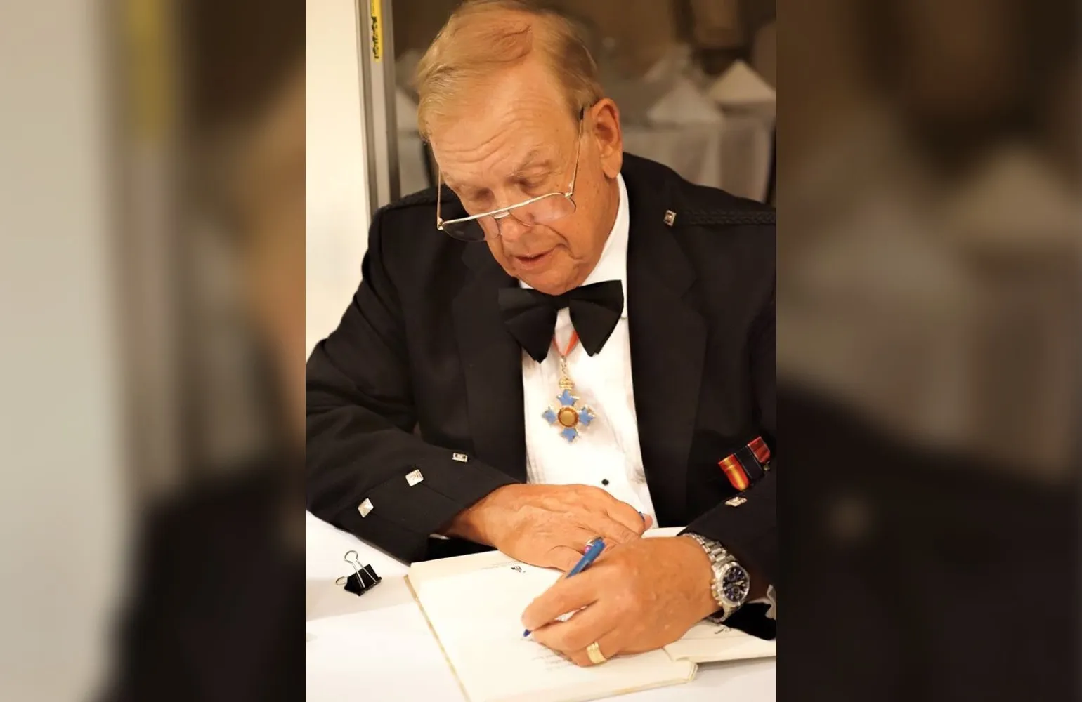 Richard M. Langworth CBE, wearing a black bow tie and jacket, sitting at a table signing one of his books.